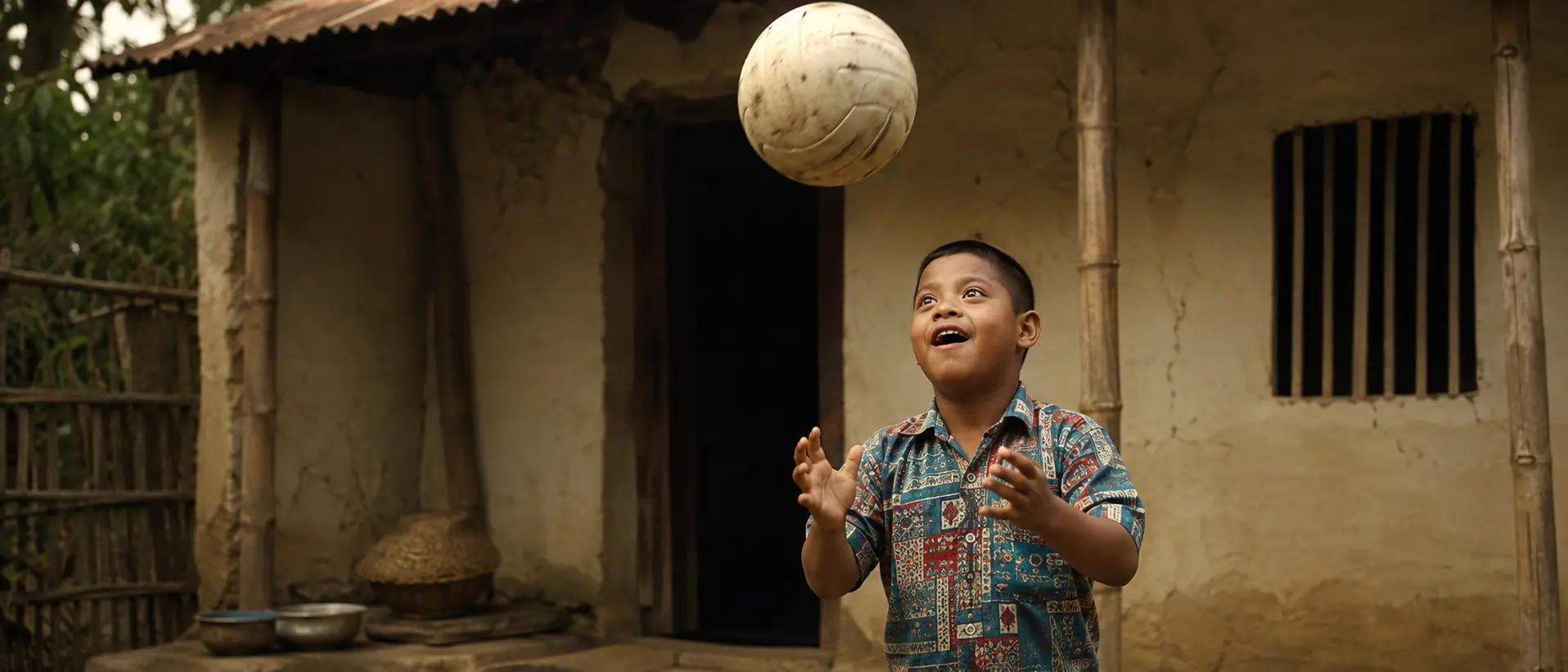 A young boy tossing a worn volleyball above his head outside his family's home, looking up at it with delight.