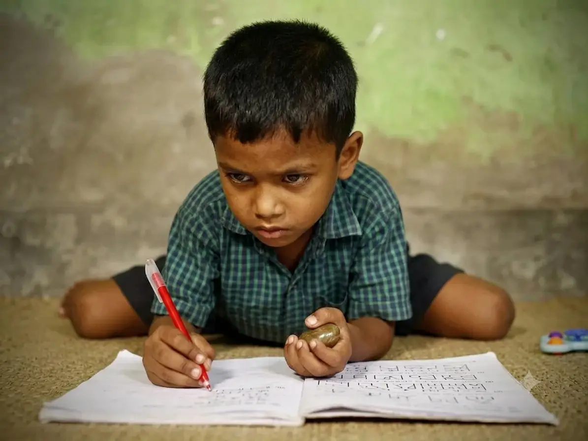 A boy lying on the floor, concentrating as he forms Bengali letters in an exercise book with a red pen.