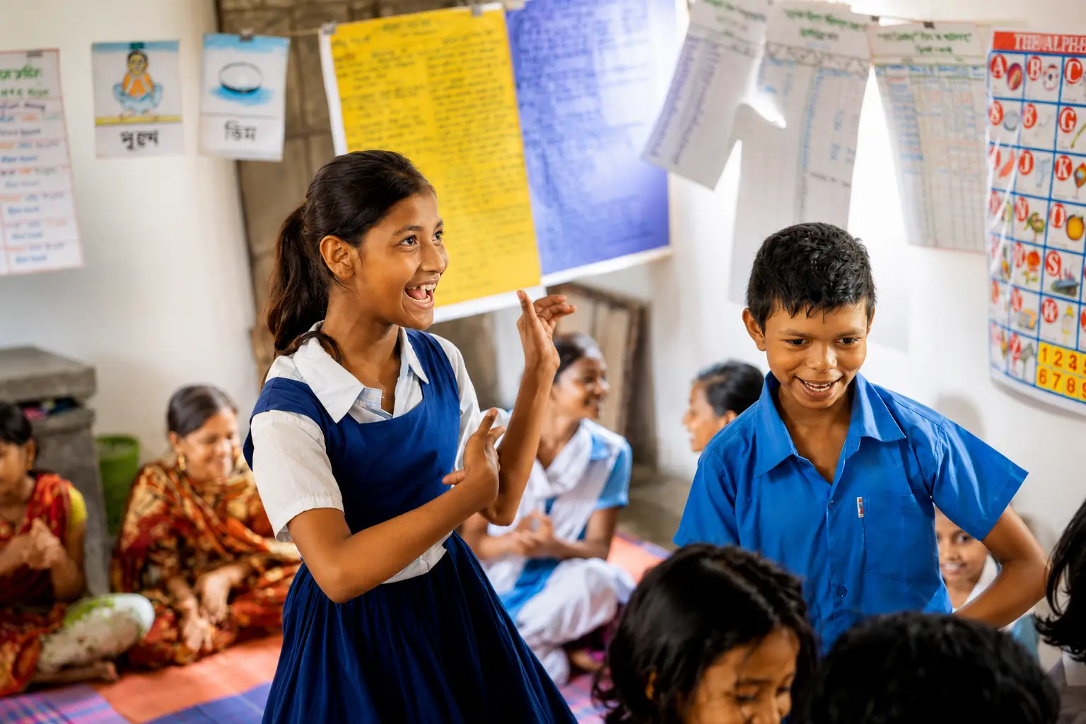 Two children laughing together in a village classroom, a girl in a school pinafore signing with her hands and a boy in a blue school shirt grinning back. Bengali and English literacy charts are pegged up across the wall behind them, and more children and women sit on a mat along the floor.