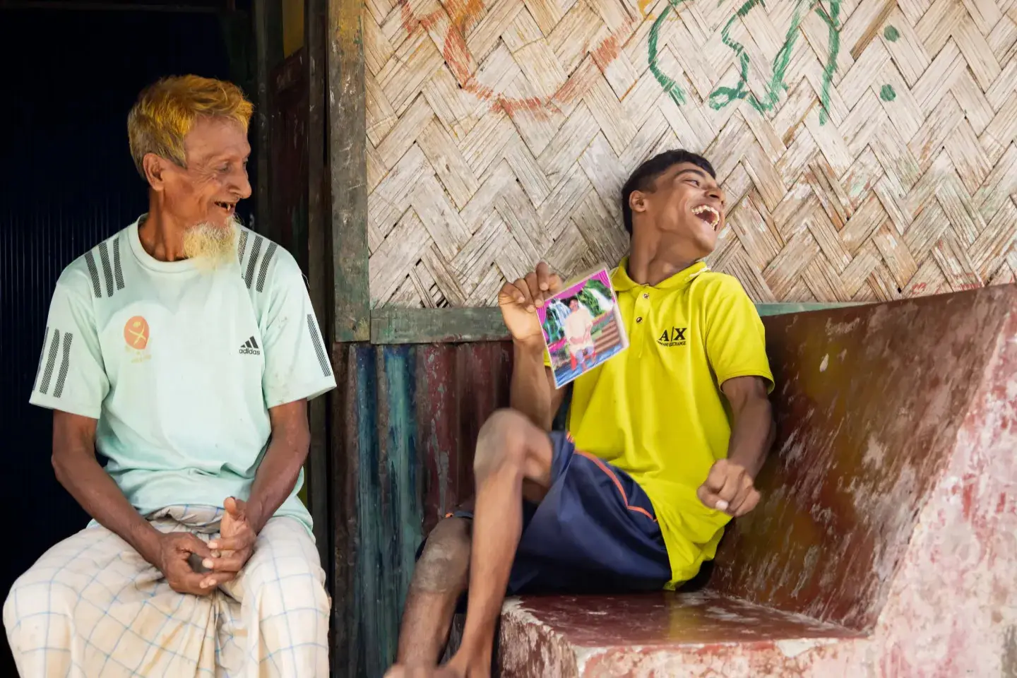 A young man in a yellow polo shirt laughing with his head tipped back against a bamboo wall, holding up a small printed photograph, while an older man in a pale green t-shirt and checked lungi sits beside him on a low step and watches him, smiling.