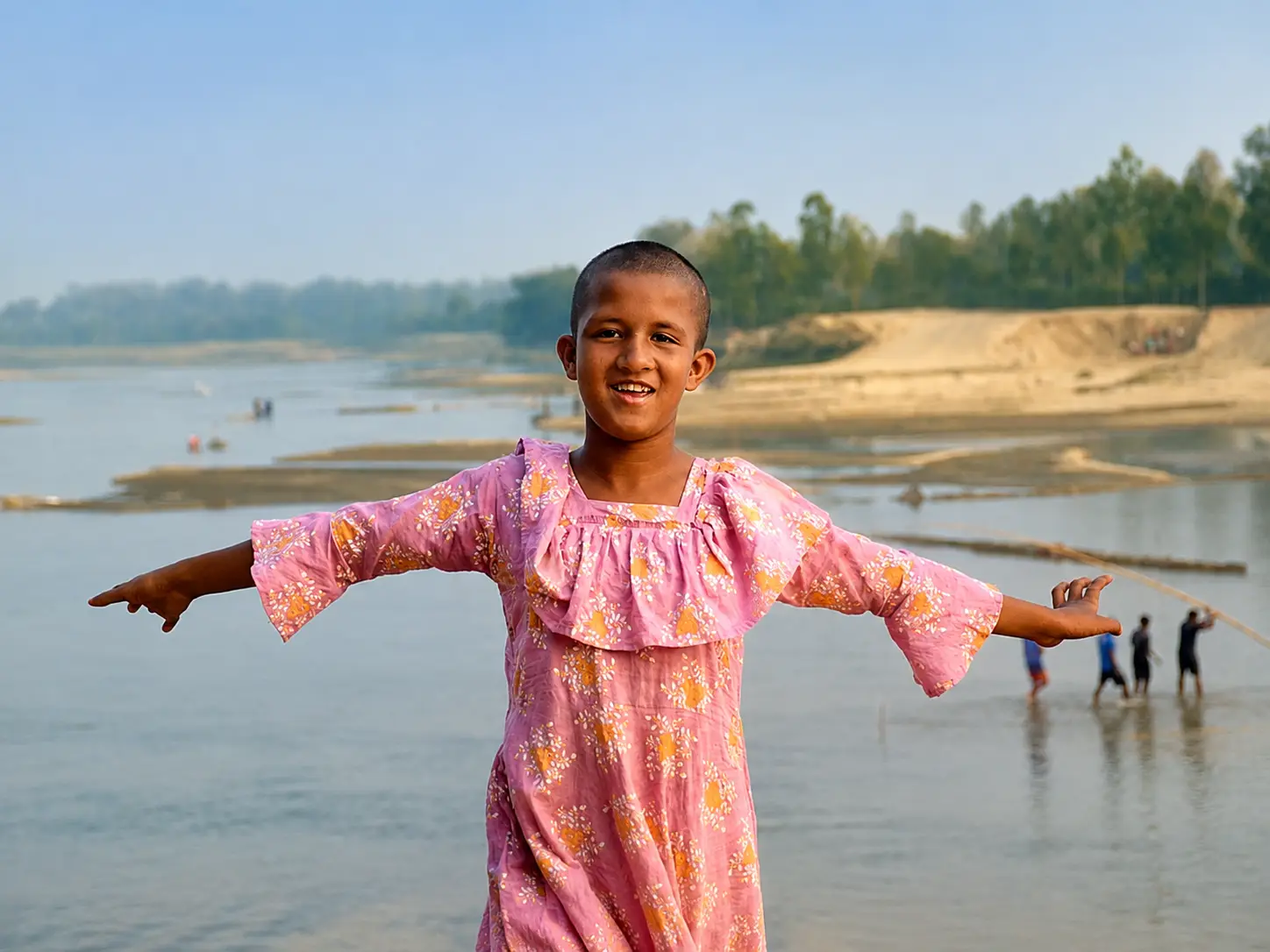 A girl standing on a wide riverbank with her arms stretched out, smiling.