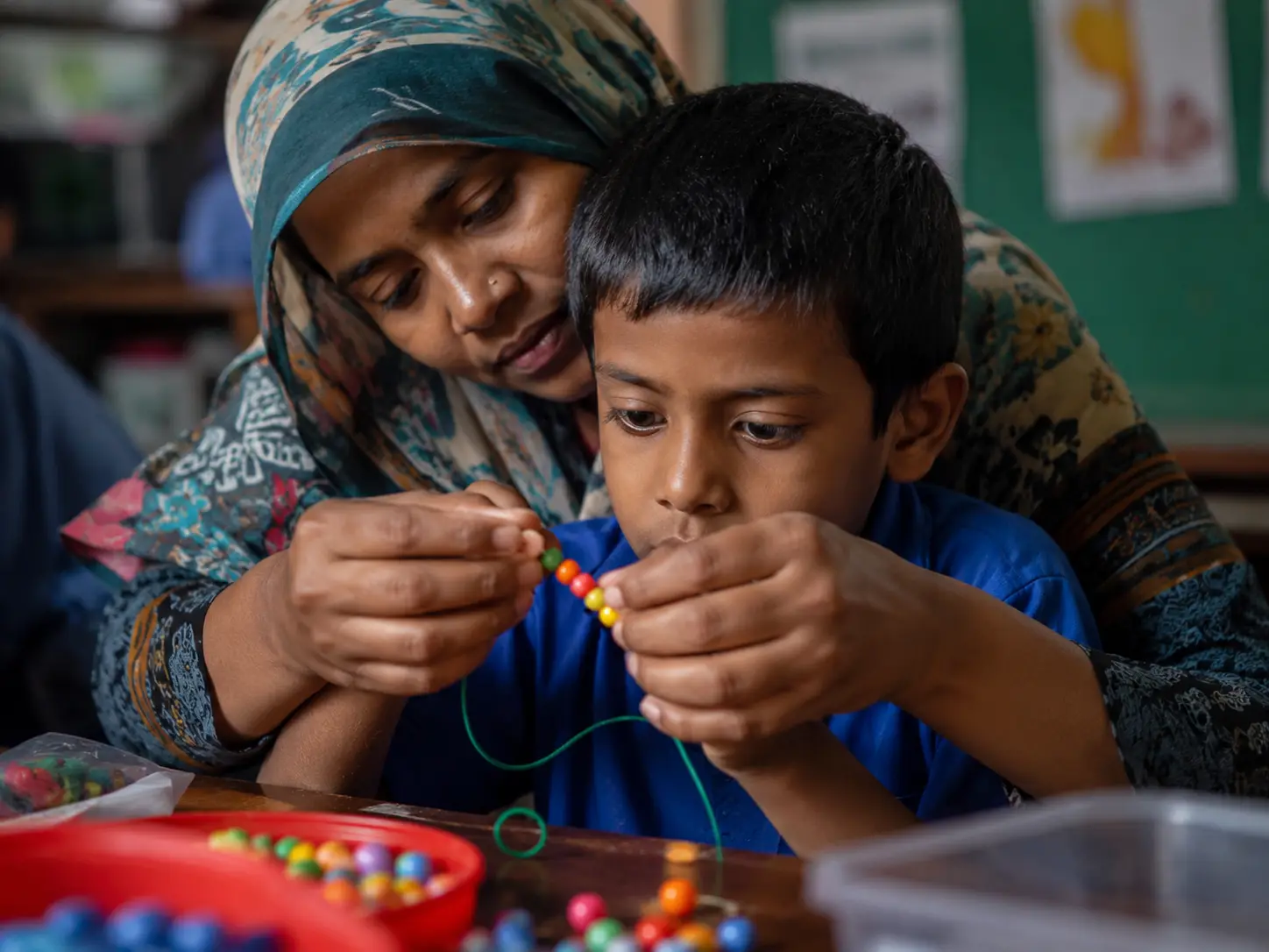 A mother guiding her son's hands as he threads coloured beads onto a green string during a therapy session.
